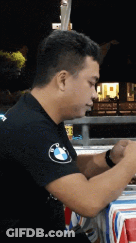 A Man Wearing A Black Bmw Shirt Sits At A Table At Night GIF