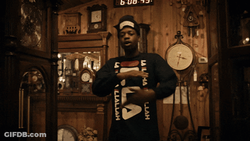 A Man Wearing A Black Fila Sweater Stands In Front Of A Wall Of Clocks GIF