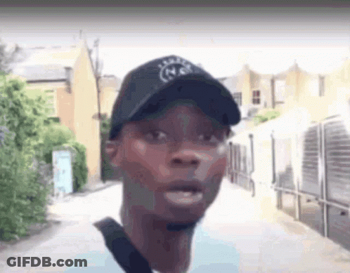 A Man Wearing A Black Hat And A Blue Shirt Is Walking Down A Street . GIF
