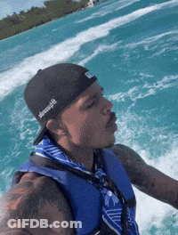 A Man Wearing A Black Hat And A Blue Vest Is Riding A Boat GIF