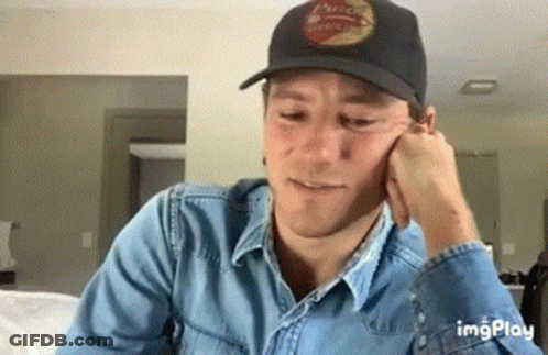 A Man Wearing A Black Hat And A Denim Shirt Is Sitting On A Couch . GIF