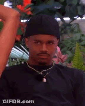 A Man Wearing A Black Hat And A Necklace Is Sitting In Front Of A Flower Garden . GIF