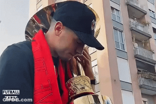 A Man Wearing A Black Hat And A Red Scarf Holds A Trophy GIF