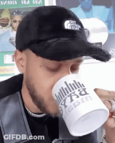 A Man Wearing A Black Hat Is Drinking From A White Mug That Says Bxa On It GIF
