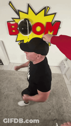 A Man Wearing A Black Hat Stands In Front Of A Boom Sign GIF