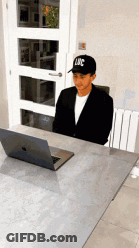 A Man Wearing A Black Hat With Luc On It Sits At A Desk GIF