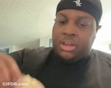 A Man Wearing A Black Hat With The Letter X On It Is Eating A Sandwich . GIF