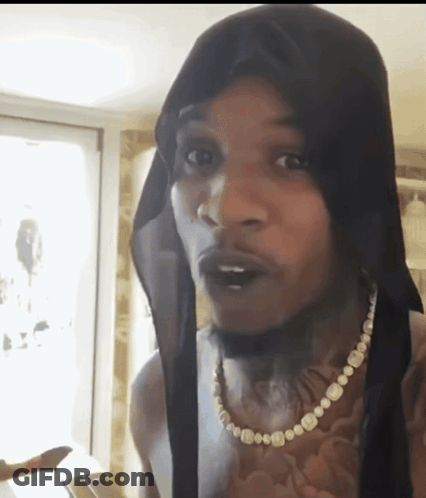 A Man Wearing A Black Head Scarf And A Pearl Necklace Looks At The Camera GIF