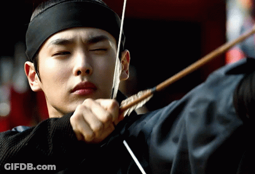 A Man Wearing A Black Headband Is Aiming A Bow And Arrow GIF