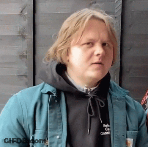 A Man Wearing A Black Hoodie And A Green Jacket Is Standing In Front Of A Wooden Fence . GIF