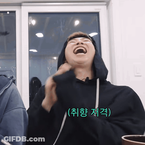 A Man Wearing A Black Hoodie Is Laughing In Front Of A Window With Korean Writing On The Bottom Right GIF