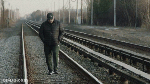 A Man Wearing A Black Jacket And A Black Hat Walks Along Train Tracks GIF
