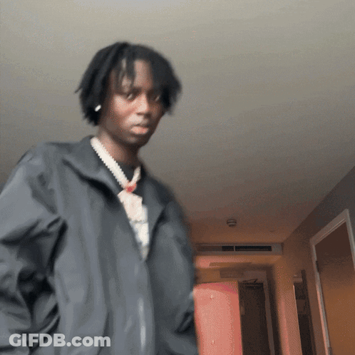 A Man Wearing A Black Jacket And A Necklace Is Standing In A Hallway GIF