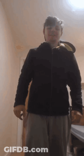 A Man Wearing A Black Jacket And Grey Pants Stands In A Hallway GIF