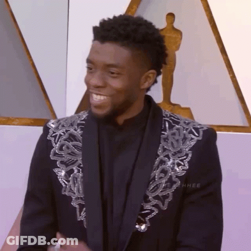 A Man Wearing A Black Jacket With Silver Sequins On The Sleeves Stands In Front Of A Statue Of An Oscar Winner GIF