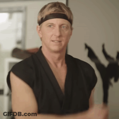 A Man Wearing A Black Karate Uniform And A Headband Is Standing In Front Of A Statue Of A Ninja . GIF