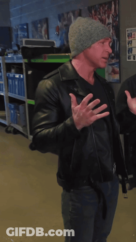 A Man Wearing A Black Leather Jacket And A Gray Hat Stands In A Room GIF