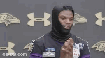 A Man Wearing A Black Mask And A Purple Jersey Is Standing In Front Of A Wall With Ravens Logos . GIF