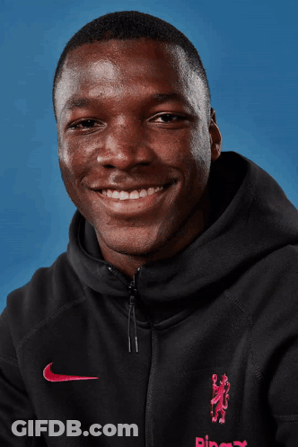 A Man Wearing A Black Nike Hoodie Smiles For The Camera Against A Blue Background GIF
