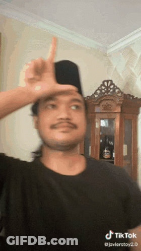 A Man Wearing A Black Shirt And A Black Hat Is Making A Funny Face With His Finger On His Forehead .. GIF