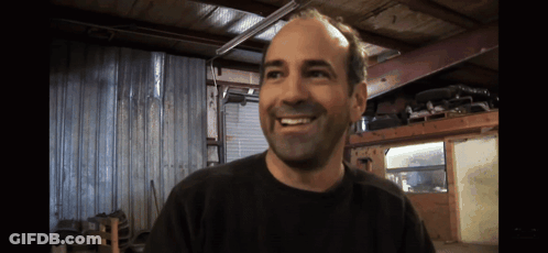 A Man Wearing A Black Shirt Is Smiling In A Garage GIF