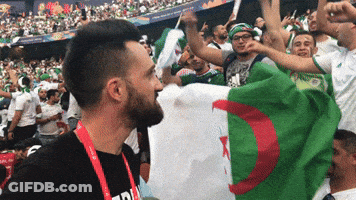 A Man Wearing A Black Shirt That Says ' A ' On It Stands In A Crowd Holding A Flag GIF