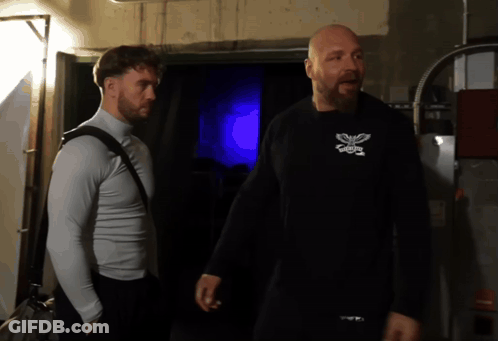 A Man Wearing A Black Shirt With A Butterfly On It Stands Next To Another Man Wearing A White Shirt GIF