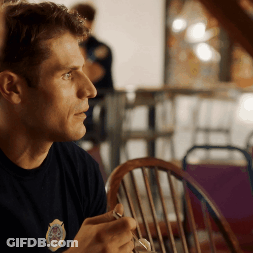 A Man Wearing A Black Shirt With A Firefighter Emblem On It Sits At A Table GIF