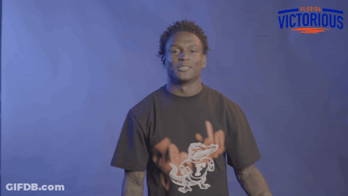 A Man Wearing A Black Shirt With An Alligator On It Stands In Front Of A Sign That Says Victorious GIF