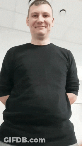 A Man Wearing A Black Shirt With His Hands In His Pockets Smiles GIF