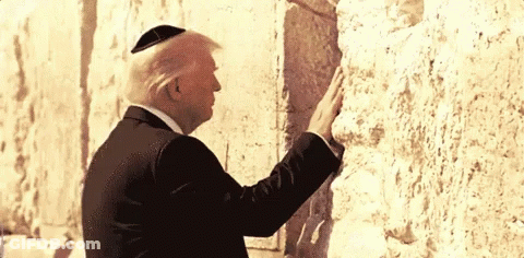 A Man Wearing A Black Suit And A Yarmulke Stands In Front Of A Stone Wall GIF