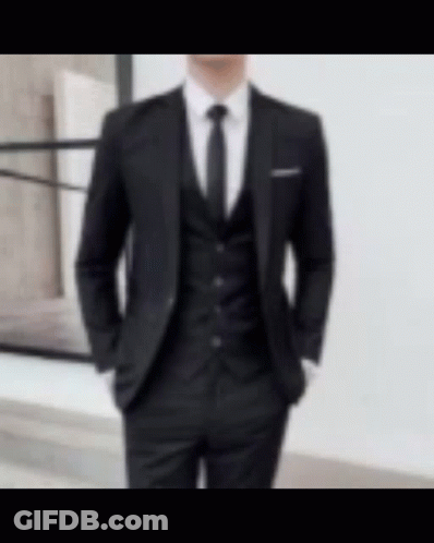 A Man Wearing A Black Suit And Tie Is Standing In Front Of A Window . GIF