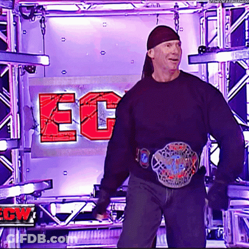 A Man Wearing A Black Sweater And A Belt Is Standing In Front Of A Sign That Says Ec On It GIF