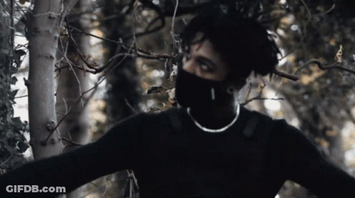 A Man Wearing A Black Sweater And A Mask Is Standing In A Forest GIF
