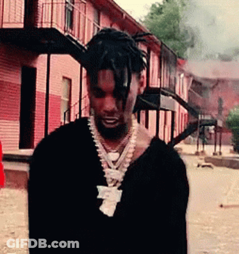 A Man Wearing A Black Sweater And A Necklace Is Standing In Front Of A Building . GIF