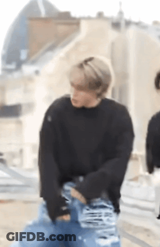 A Man Wearing A Black Sweater And Blue Jeans Is Standing On A Sidewalk . GIF