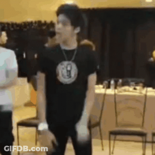 A Man Wearing A Black T-Shirt And A Necklace Is Standing In A Room . GIF