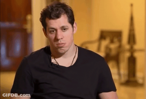 A Man Wearing A Black T-Shirt Is Sitting In Front Of A Chair GIF