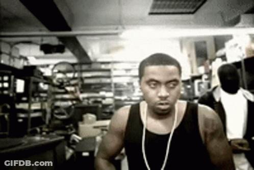 A Man Wearing A Black Tank Top And A Necklace Stands In A Room GIF