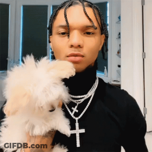 A Man Wearing A Black Turtleneck And A Necklace With A Cross On It Holds A Small White Dog GIF