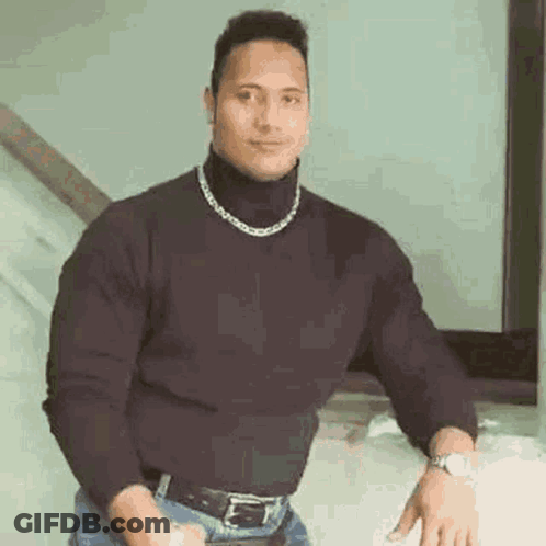 A Man Wearing A Black Turtleneck Sweater And A Chain Around His Neck Is Standing In Front Of A Mirror . GIF