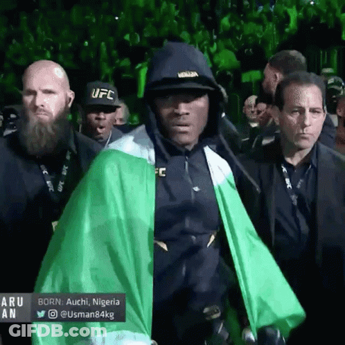 A Man Wearing A Black Ufc Hat And A Green Cape Walks Through A Crowd GIF