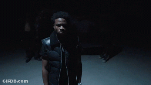 A Man Wearing A Black Vest And Headphones Stands In The Dark GIF