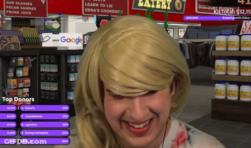 A Man Wearing A Blonde Wig Is Smiling In Front Of A Sign That Says Eatery GIF