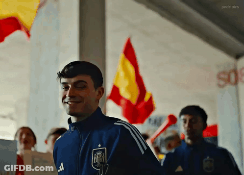 A Man Wearing A Blue Adidas Jacket Is Smiling In Front Of A Group Of People Holding Spanish Flags . GIF