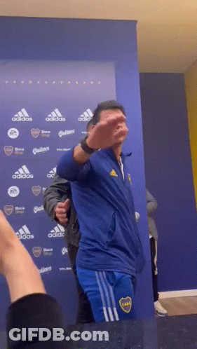 A Man Wearing A Blue Adidas Jacket Stands In Front Of A Blue Wall GIF