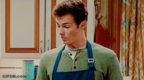 A Man Wearing A Blue Apron Is Standing In A Kitchen . GIF