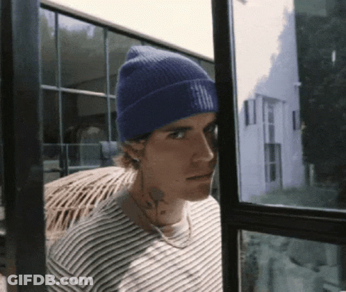 A Man Wearing A Blue Beanie And A Striped Shirt Looks Out Of A Window GIF