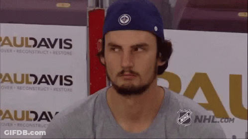 A Man Wearing A Blue Hat And A Grey Shirt Is Sitting In Front Of A Sign That Says Audavis . GIF