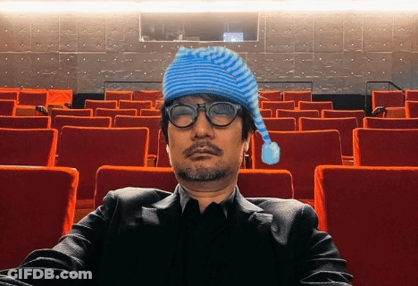 A Man Wearing A Blue Hat And Glasses Sits In A Theater GIF